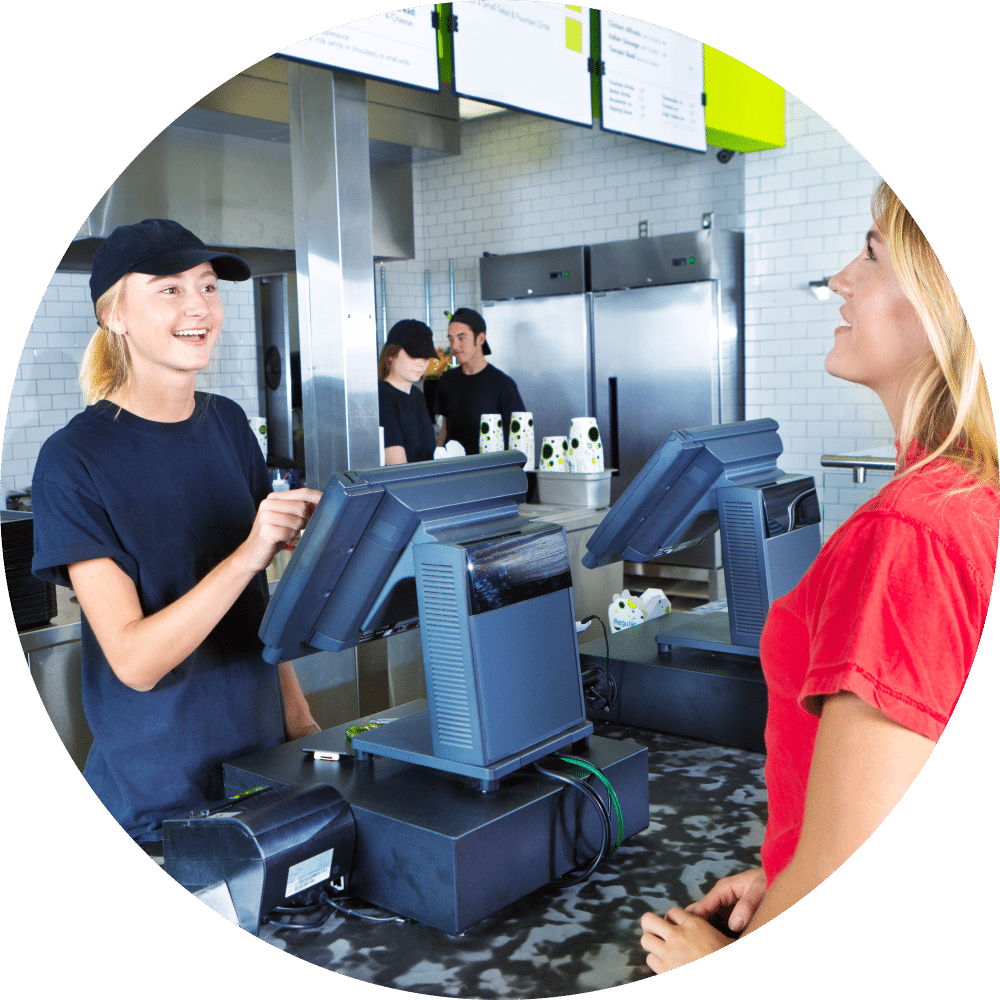 A young woman customer placing her order at a fast food convenience restaurant. A young woman server staff is assisting her at the checkout cashier counter with the kitchen staff working in the background.